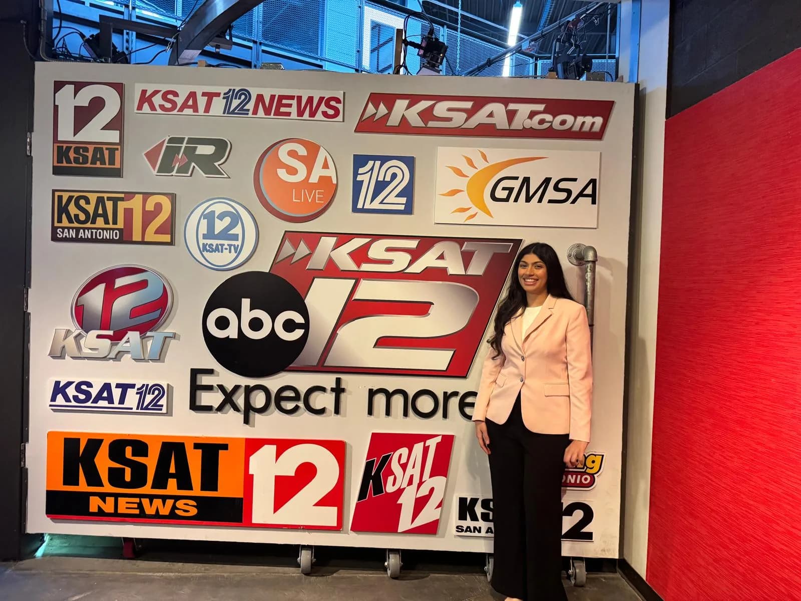 Nikita Tejwani smiling, in a pink blazer, standing beside a wall of KSAT 12 station logos at the KSAT studio.