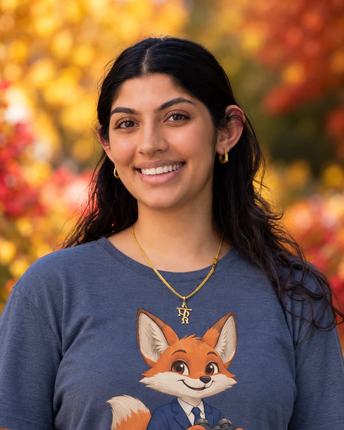 Close portrait of Nikita Tejwani smiling, in a navy t-shirt with the Caly fox on it, in front of autumn leaves.