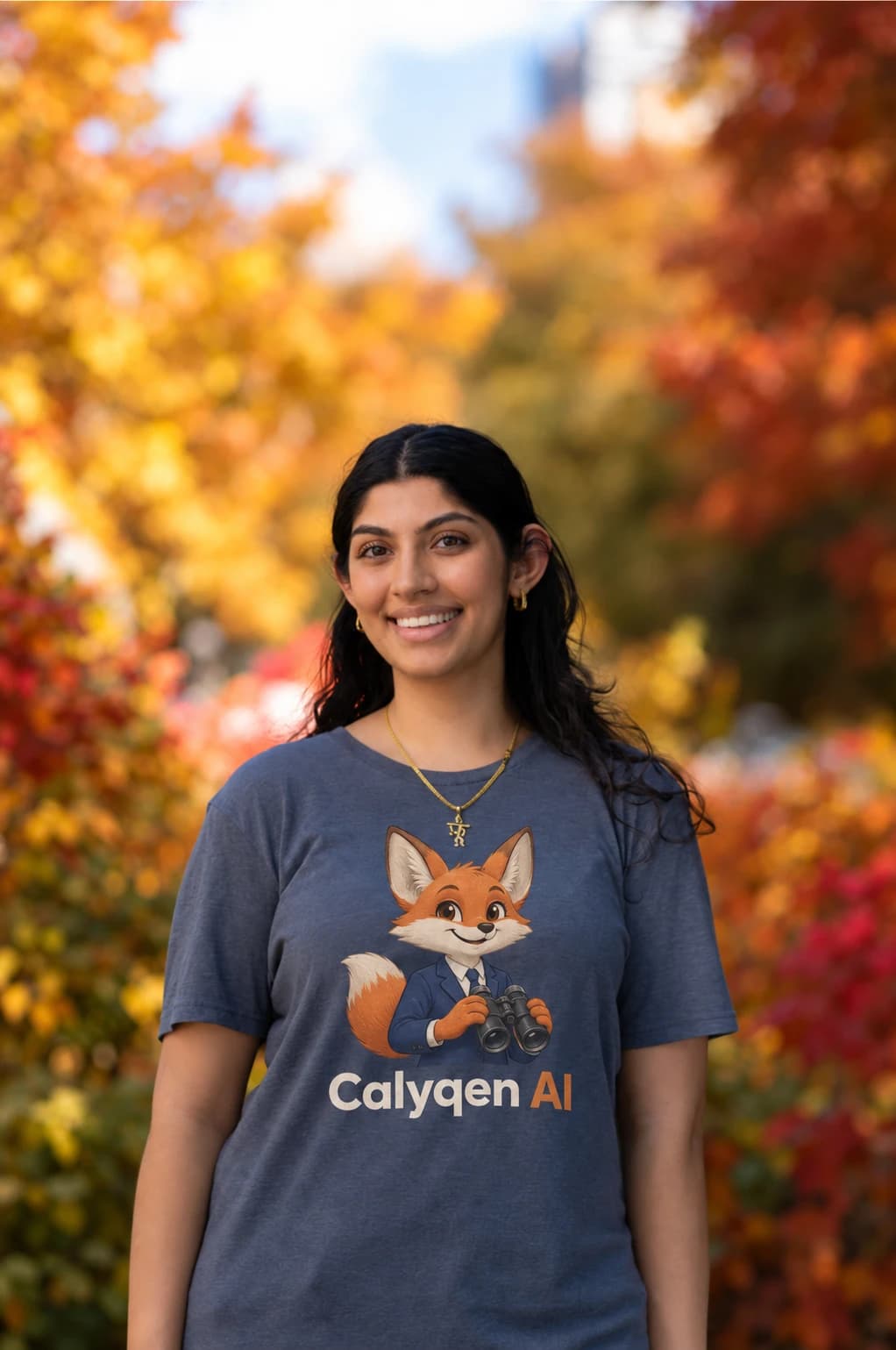 Nikita Tejwani standing and smiling, in a navy Calyqen AI t-shirt showing the Caly fox holding binoculars, in front of autumn leaves.