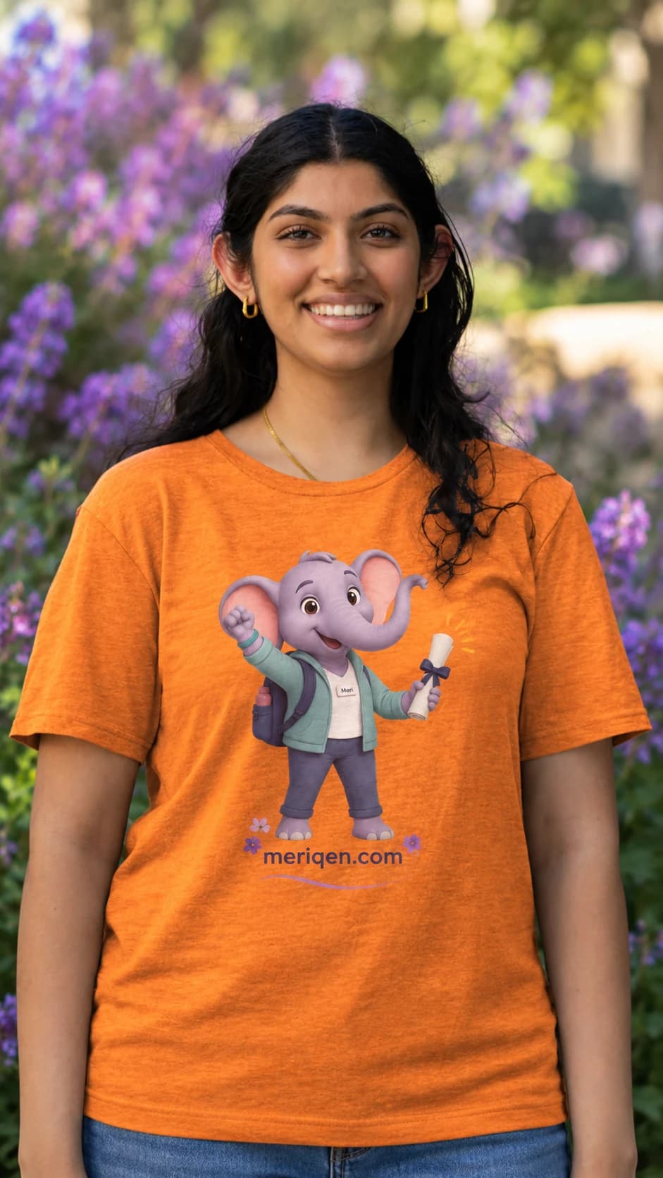 Nikita Tejwani smiling, in an orange t-shirt showing Meri the elephant holding a rolled diploma, with meriqen.com beneath, in front of purple flowers.