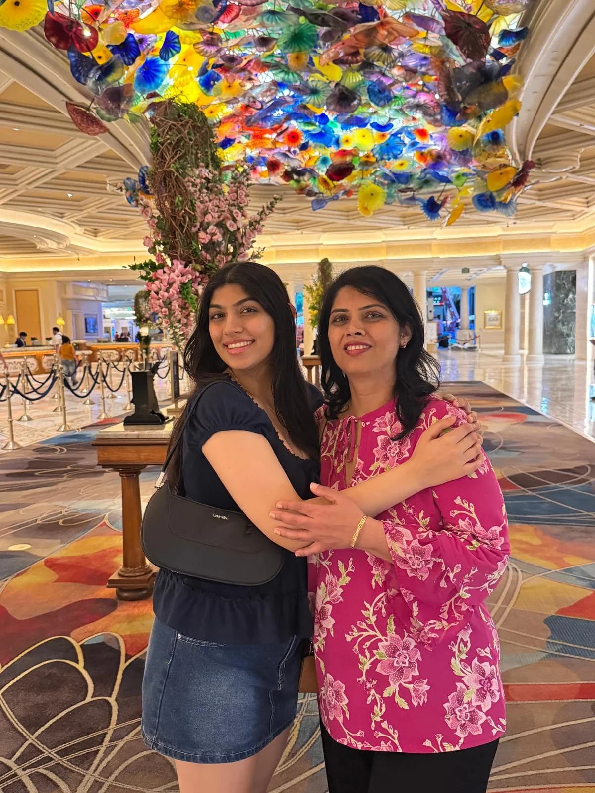 Nikita and her mom hugging and smiling in a hotel lobby, under a ceiling of colourful glass flowers.