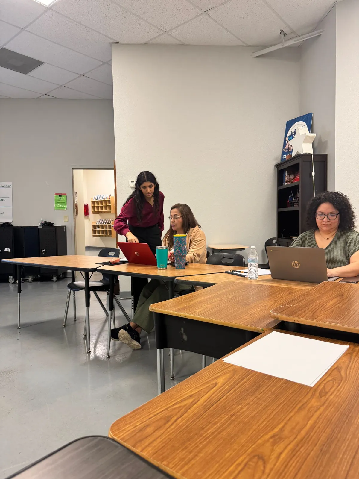 Nikita leaning over a red laptop to show an SA Youth staff member something on the screen, with another staff member working at a desk beside them.