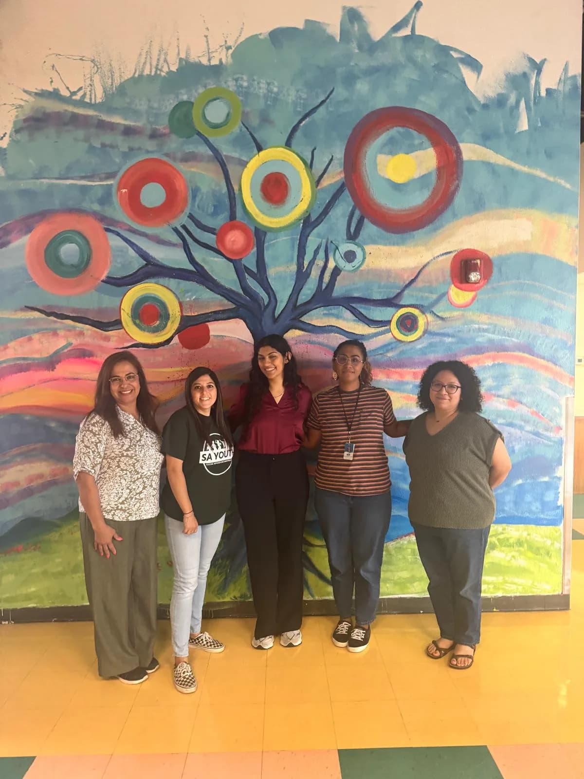 Nikita standing with four people from SA Youth in front of a colourful painted mural of a tree, all smiling.