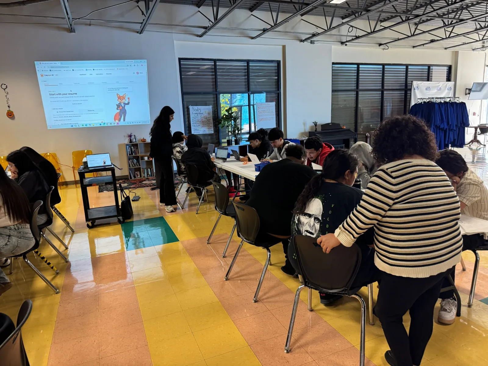 Students working on laptops at long tables while Nikita checks in near the projector screen.