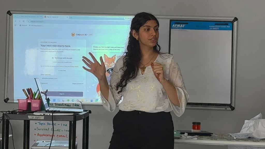 Nikita speaking in an SA Youth classroom, gesturing as she explains, with Calyqen's sign-in screen projected behind her.