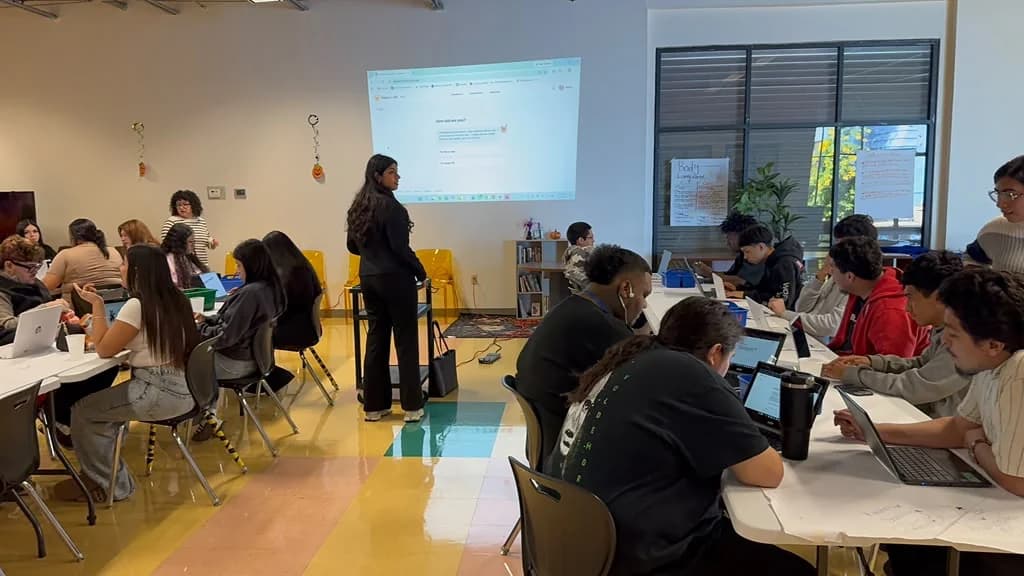 Nikita stands beside a projector screen showing a Calyqen question while students work at tables with laptops.
